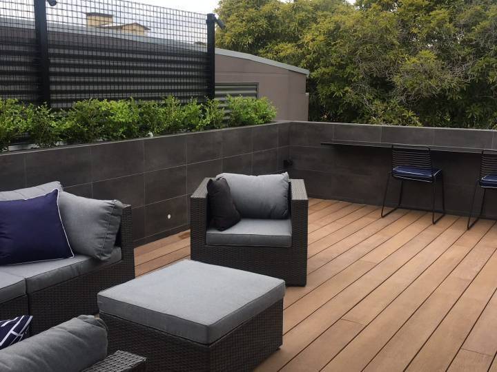 Modern rooftop terrace featuring sleek wood decking, comfortable outdoor seating with plush cushions, and a minimalist bar area with built-in stools, surrounded by greenery and privacy walls.