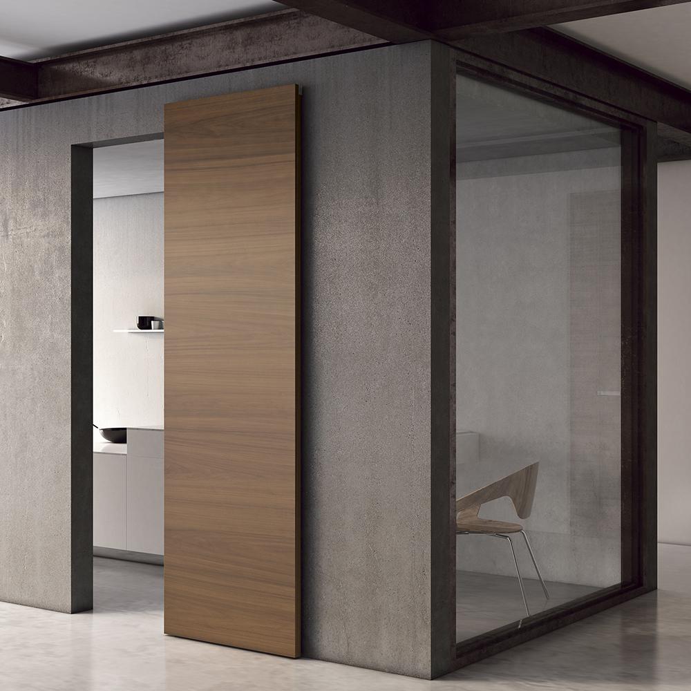 MODERN_SLIDING_DOORS_FILA_AMERICAN_WALNUT_NATURE Minimalist interior with a wooden sliding door mounted on a dark frame, adjacent to a glass-walled office space.