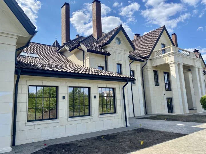 A newly constructed elegant house featuring classical architectural elements, cream-colored stone façade, dark-framed windows, and a tiled roof with multiple chimneys.