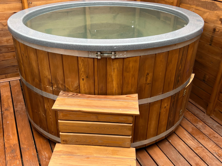 A round wooden hot tub filled with water, featuring a sturdy built-in step for easy access, placed on a timber deck within a cozy enclosed space.