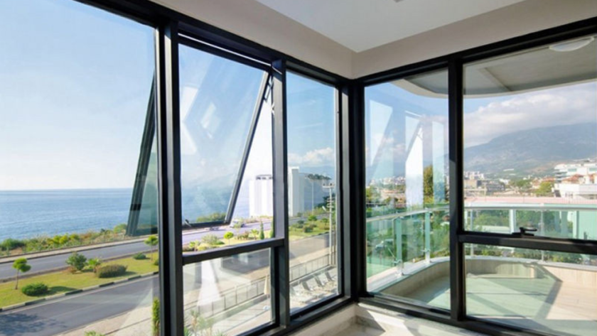View through modern European aluminium windows in a corner room, overlooking a coastal road, sea, and distant mountains under a bright sky