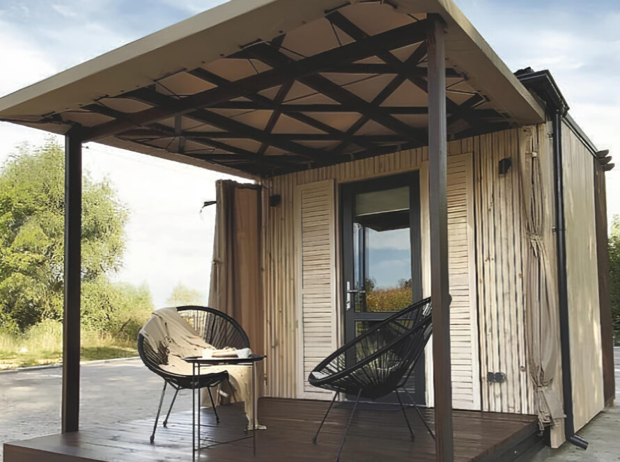 Prefab tiny guest house with covered patio, two modern chairs, and a table, set outdoors on a concrete surface with trees in the background