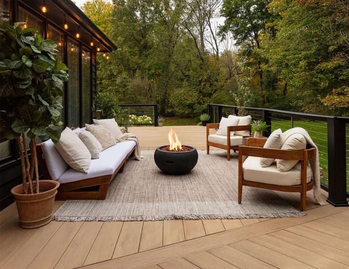 A stylish outdoor living space with warm-toned wood-effect decking, modern lounge seating, a fire pit centerpiece, and surrounded by lush greenery.