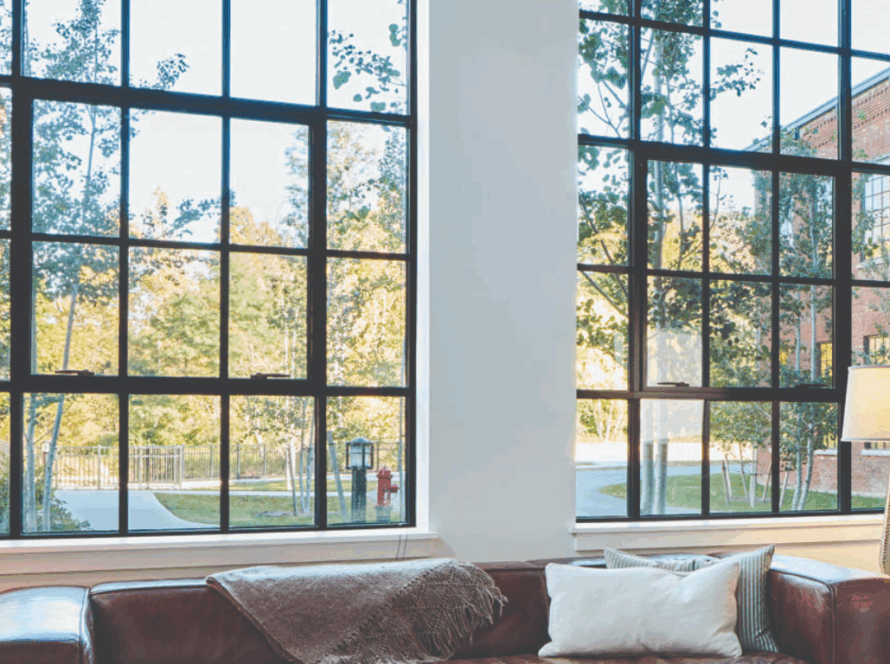 Black steel casement windows with grid pattern in a living room, offering a clear view of trees and buildings outside, paired with a leather sofa and lamp