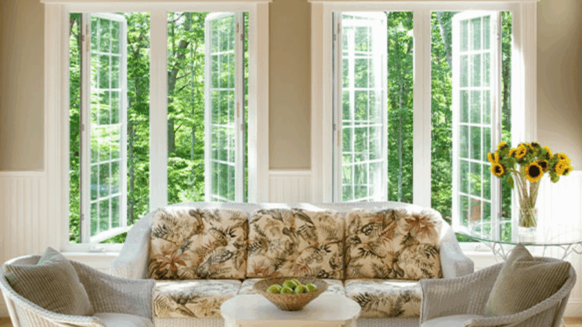 Bright living room with large white vinyl casement windows opened outward, surrounded by greenery, sofa and sunflowers in foreground