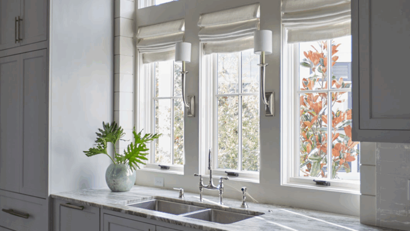 White casement windows above a double kitchen sink, framed by light-colored walls and Roman shades, offering a view of autumn foliage outside
