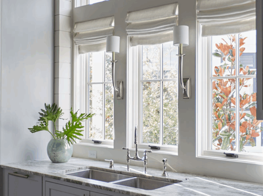 White casement windows above a double kitchen sink, framed by light-colored walls and Roman shades, offering a view of autumn foliage outside