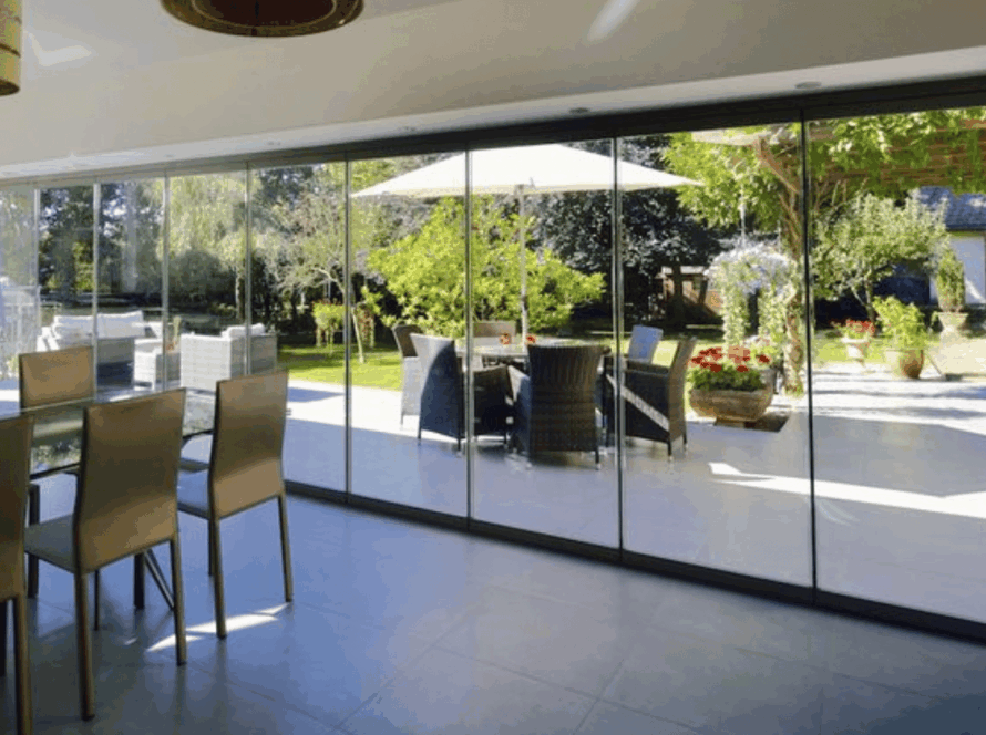 Frameless sliding glass window overlooking a garden patio with outdoor seating and greenery