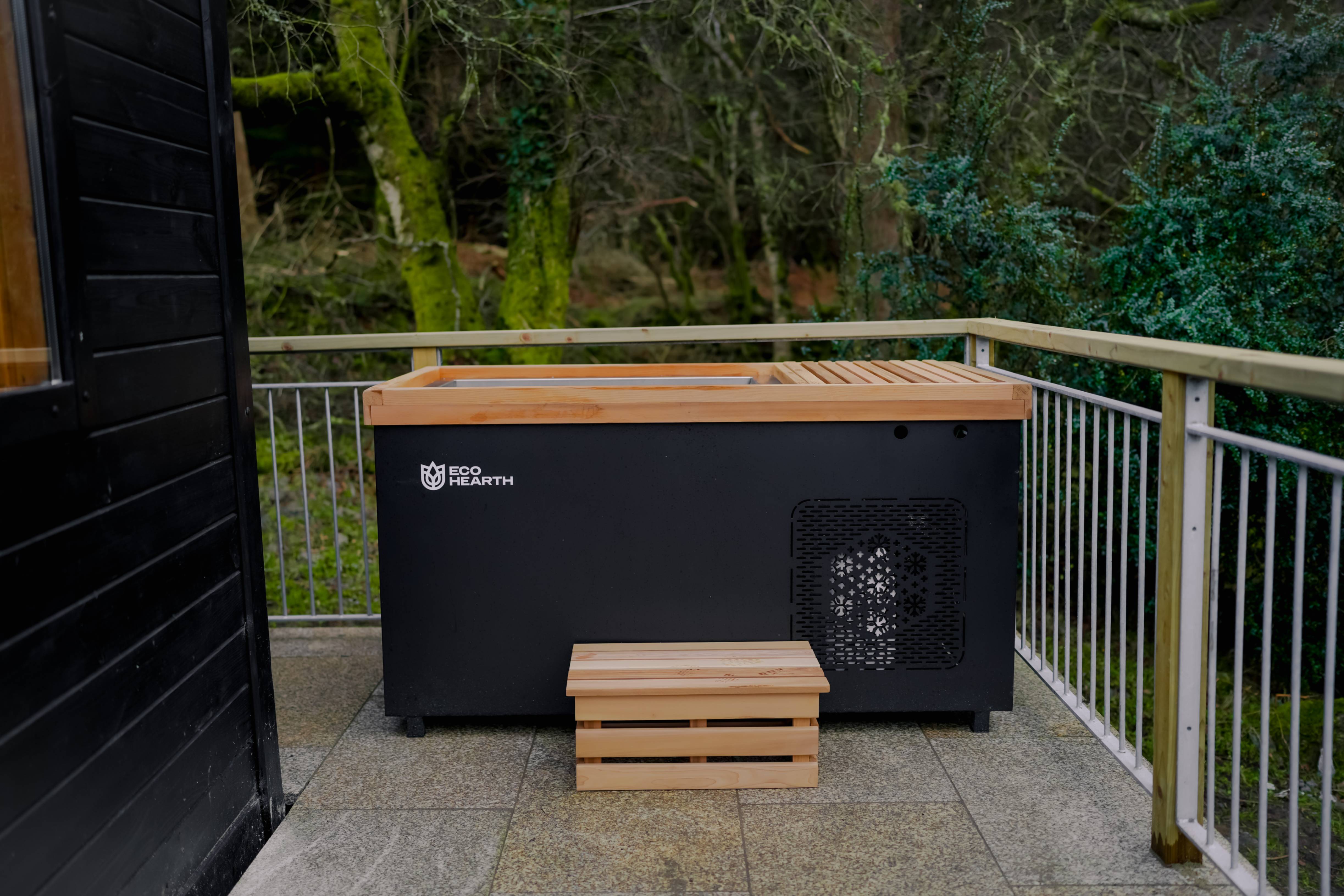 Eco Hearth outdoor ice bath tub with wooden lid and step, modern black cold plunge installed on a terrace surrounded by nature