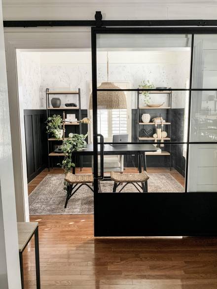 A stylish interior space featuring a black-framed lift & slide door with glass panels, separating a cozy, modern office or dining area with natural wood flooring and minimalistic decor.