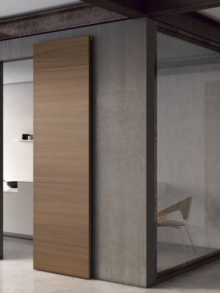 MODERN_SLIDING_DOORS_FILA_AMERICAN_WALNUT_NATURE Minimalist interior with a wooden sliding door mounted on a dark frame, adjacent to a glass-walled office space.